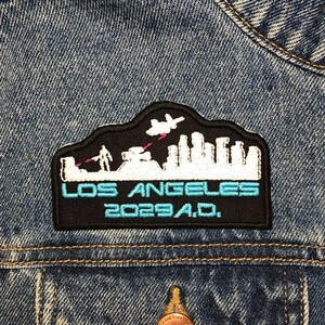 Może przedstawiać: Naszywkę haftowana w kolorze czarno-białym z panoramą miasta i tekstem "LOS ANGELES 2023 A.D." Naszywka jest przyszyta do niebieskiej kurtki dżinsowej.