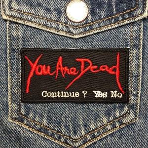 May include: A black denim jacket with a black patch that says "You Are Dead" in red lettering. The patch also says "Continue? Yes No" in white lettering.