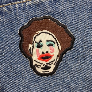 Op de afbeelding: Een geborduurde patch van een gezicht met een clown-achtige make-up, met bruin haar en een witte huid. De patch is genaaid op een blauwe denim achtergrond.