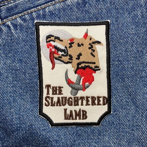 May include: A white patch with a brown embroidered image of a wolf's head with a hook and blood dripping from its mouth. The text "THE SLAUGHTERED LAMB" is embroidered in brown below the image.
