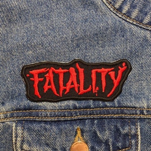May include: A blue denim jacket with a black and red patch. The patch, sewn onto the jacket, displays the word "FATALITY" in red, stylized lettering. The jacket has a chest pocket with a copper-coloured button.
