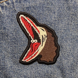 Peut inclure: Un patch brodé représentant un monstre de dessin animé avec une grande bouche ouverte et des cheveux bruns, cousu sur une veste en jean bleue. Le patch est rouge et blanc.
