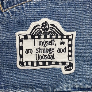May include: A white embroidered patch with black stitching on a blue denim jacket. The patch features a black and white striped border and the text "I myself, am strange and unusual."