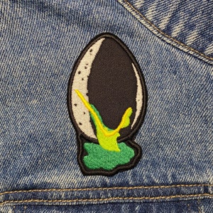 May include: An embroidered patch of an alien creature from the film 'Alien' on a blue denim jacket. The patch is black, green and yellow.
