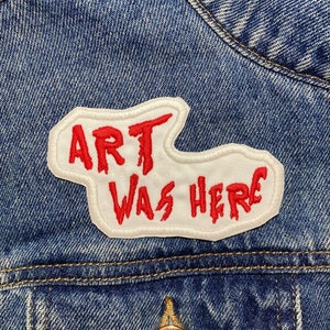 May include: A white patch sewn onto a blue denim jacket with red embroidered text that reads "ART WAS HERE".