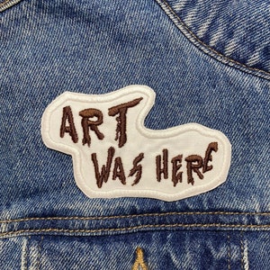 以下が含まれることがあります： 青いデニムジャケットに、茶色のステッチが施された白いパッチ。パッチには「Art Was Here.」と書かれている。