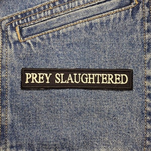 Może przedstawiać: Zbliżenie na niebieską dżinsową kurtkę z czarną naszywką, na której widnieje napis "PREY SLAUGHTERED" żółtymi literami.