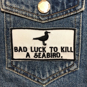 May include: A white patch sewn onto a blue denim jacket with black stitching. The patch features a black silhouette of a seabird and the text "BAD LUCK TO KILL A SEABIRD."