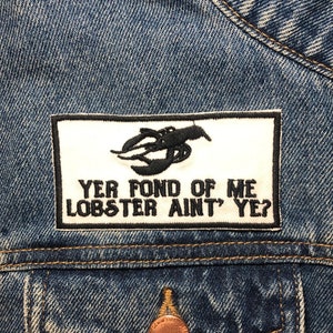 Könnte beinhalten: Ein weißer Patch, der auf eine blaue Jeansjacke mit schwarzer Stickerei eines Hummers und dem Text "Yer fond of me lobster ain't ye?" genäht ist.