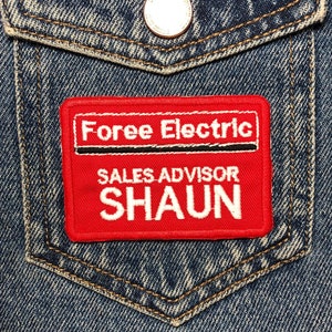 Peut inclure: Gros plan d'une poche de veste en jean avec un écusson rouge. L'écusson indique "Foree Electric", "SALES ADVISOR" et "SHAUN" en blanc. La veste est en jean bleu avec un bouton.