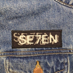 以下が含まれることがあります： 白い文字で「SE7EN」と書かれた黒いパッチが付いたデニムジャケットです。