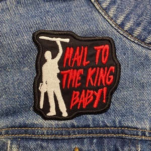 May include: A black and red embroidered patch with a white silhouette of a person holding a chainsaw. The text on the patch reads "Mail to the King Baby!"