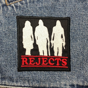 Może przedstawiać: Czarno-biała łatka z trzema sylwetkami i napisem "REJECTS" czerwonymi literami.