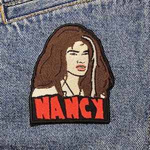 May include: A brown and black embroidered patch with a cartoon image of a woman with brown hair and the word "NANCY" in red letters.