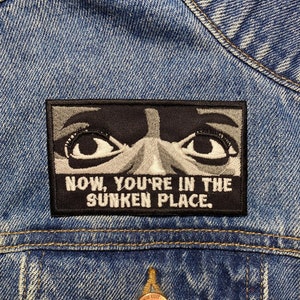 Op de afbeelding: Een zwart-witte patch met een paar ogen en de tekst "NOW, YOU'RE IN THE SUNKEN PLACE."