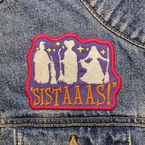 Pode incluir: Patch bordado roxo e rosa com três figuras brancas segurando vassouras e o texto "Sistaas!" em letras laranja.