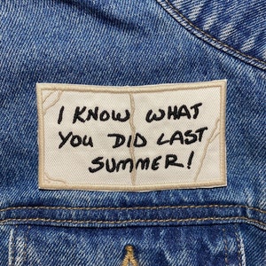Peut inclure: Un écusson blanc cousu sur une veste en jean bleue avec le texte "I know what you did last summer!" écrit en noir.