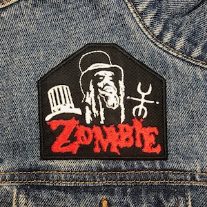 Może przedstawiać: Czarno-czerwona naszywka haftowana z napisem "Zombie" w czerwonych literach. Naszywka przedstawia białą sylwetkę osoby z długimi włosami w kapeluszu i białym symbolem.