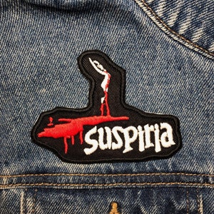 May include: Black and red embroidered patch with the word "Suspiria" in white lettering. The patch features a silhouette of a person with red blood dripping from their hands.