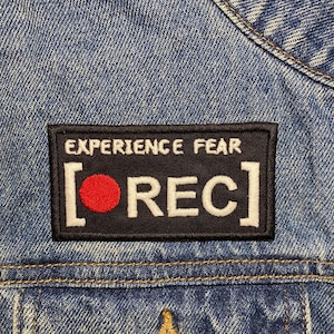May include: Black patch with white text that reads "EXPERIENCE FEAR [REC]" with a red circle inside the brackets. The patch is sewn onto a blue denim jacket.