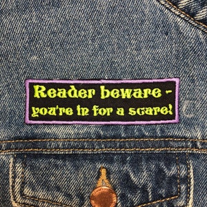 以下が含まれることがあります： 「Reader beware - you're in for a scare!」と書かれた紫と黒の刺繍パッチが付いた青いデニムジャケット