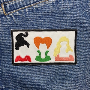 Könnte beinhalten: Ein weißer Patch mit schwarzem Rand, der vier bunte gestickte Silhouetten von Hexen zeigt. Die Hexen tragen rote, grüne und rosa Outfits mit orangefarbenem, gelbem und weißem Haar.