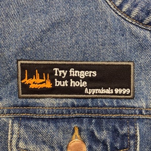 Może przedstawiać: Czarna kurtka dżinsowa z naszywką, na której widnieje napis "Try fingers but hole" i "Appraisals 9999" w pomarańczowym kolorze.