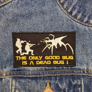 May include: A black and yellow embroidered patch on a blue denim jacket. The patch features a white silhouette of a bug with multiple legs and two figures shooting at it. The text on the patch reads "The only good bug is a dead bug!"