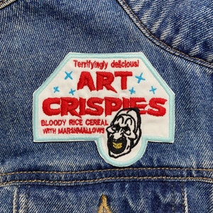 Può includere: Una toppa ricamata bianca e rossa con il testo "Terrifiyingly delicious! Art Crispies Bloody Rice Cereal with Marshmallows" e un'immagine di un clown con un sorriso pieno di denti.