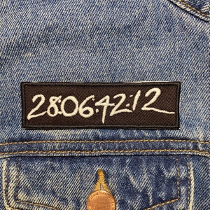 Op de afbeelding: Een zwarte patch met witte stiksels op een blauwe denimjas. De patch heeft de tekst "28:06:42:12" erop.