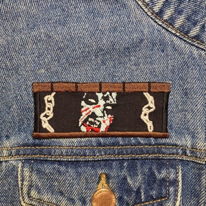 May include: A black and white embroidered patch featuring a skull and crossbones design. The patch is bordered in brown with a chain link design.