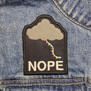 May include: A black and grey embroidered patch with a grey cloud and a colourful string of lights hanging from it. The patch says "NOPE" in white letters.
