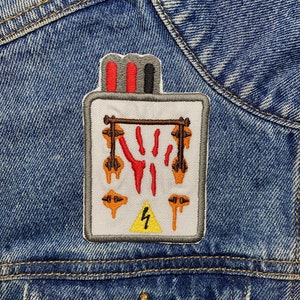 May include: A white embroidered patch with a red, black, and gray fuse box design. The fuse box has a yellow lightning bolt warning symbol and orange dripping wires.