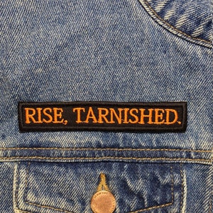 Könnte beinhalten: Eine schwarze Jeansjacke mit einem schwarzen Patch, auf dem "RISE, TARNISHED." in orangefarbener Schrift steht.