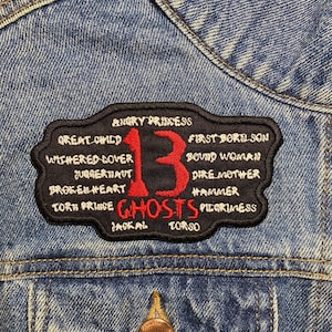 May include: A black and red embroidered patch with the number "13" in red and the word "GHOSTS" in black. The patch is surrounded by a black border and has various words in white text around the border, including "Angry Princess", "Great Child", "First Borilson", "Withered Dover", "Juggernaut", "Broken Heart", "Torn Prince", "Jackal", "Bound Woman", "Dire Mother", "Hammer", "Pilgrimess", and "Torso".