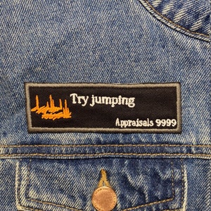 以下が含まれることがあります： 青いデニムジャケットに、「Try Jumping」と「Appraisals 9999」の文字が書かれた黒とオレンジの刺繍パッチが付いている。