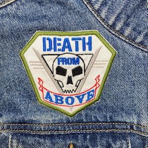 Può includere: Una toppa ricamata verde, bianca e rossa con un teschio e ossa incrociate e il testo "DEATH FROM ABOVE".