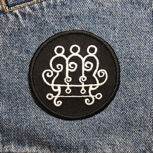 Può includere: Una toppa ricamata in bianco e nero con un simbolo bianco su sfondo nero. Il simbolo è un cerchio con una serie di volute e linee all'interno.
