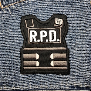 May include: Black and white embroidered patch of a bulletproof vest with the text "R.P.D." on a blue denim background.