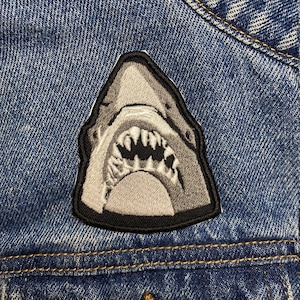 May include: A white and grey shark head patch with black stitching, sewn onto a blue denim jacket.