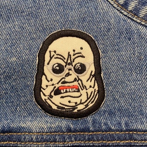 May include: A white and black embroidered patch of a monster's face with a stitched-on black border. The monster has a large, open mouth with red lips and a white tongue. The monster has large, black eyes and a white face with black lines.