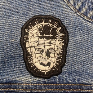 May include: A black and white embroidered patch of a character from the horror movie "Saw". The character has a barbed wire mask and a distressed look on their face. The patch is sewn onto a blue denim jacket.