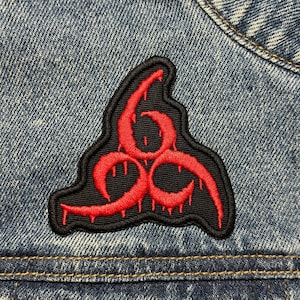 Könnte beinhalten: Roter und schwarzer gestickter Aufnäher mit einem Biohazard-Symbol mit tropfendem Blut. Der Aufnäher ist auf eine blaue Jeansjacke genäht.