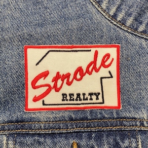 May include: A white and red embroidered patch with the text "Strode Realty" on a blue denim jacket.