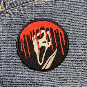 Può includere: Una toppa ricamata in bianco e nero con un design rosso che cola. La toppa presenta una figura spettrale con un coltello. La toppa è cucita su una giacca di jeans blu.