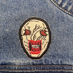 Op de afbeelding: Een rood en wit geborduurde patch van een eng gezicht met scherpe tanden en rode ogen op een blauw denim jack.