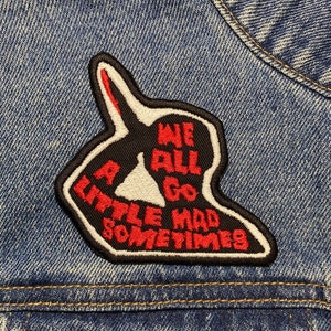 May include: An iron-on patch made of black and white fabric with red stitching. The patch shows a hand giving the middle finger with the text "We all go a little mad sometimes".