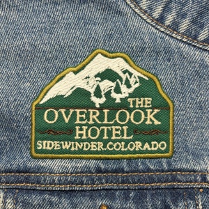 Peut inclure: Un écusson brodé vert et blanc avec le texte "The Overlook Hotel Sidewinder Colorado" sur une veste en jean bleue.