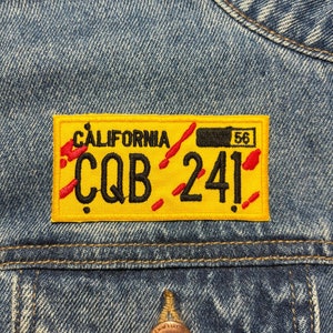 Op de afbeelding: Geel Californië kenteken patch met rode schuine strepen en zwarte tekst die "CQ B 24!" en "56" in de rechterbovenhoek leest.