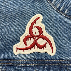 Może przedstawiać: Czerwona haftowana naszywka z kropelkowym symbolem na białym tle. Symbol to stylizowana liczba 6 z trzema pętlami.
